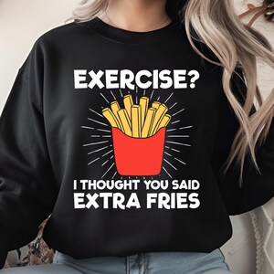 Puede incluir: Sudadera negra con el texto "EXERCISE? I THOUGHT YOU SAID EXTRA FRIES" en blanco. El gráfico presenta un recipiente rojo de patatas fritas amarillas con líneas blancas radiantes.
