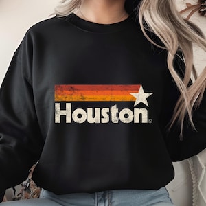 May include: Black sweatshirt featuring the word "Houston" in a distressed, vintage-style font. Above the text is a horizontal stripe design in shades of orange and red, with a white star on the right side.