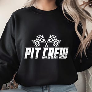 Może przedstawiać: Czarna bluza z białym napisem "PIT CREW" i dwoma skrzyżowanymi flagami w szachownicę. Projekt jest w stylu vintage, co nadaje mu wygląd retro. Bluza ma okrągły dekolt.