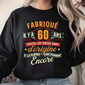 May include: Black sweatshirt with the text "FABRIQUÉ ILYA 60 ANS TOUTES LES PIECES SONT d'origine ET LA PLUPART FONCTIONNENT Encore" in orange, red, and yellow. The text is arranged in a diamond shape with stars.