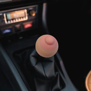 Op de afbeelding: Een novelty versnellingspookknop in de vorm van een borst, met een roze tepel en tepelhof. De knop is gemonteerd op een zwarte leren versnellingspookhoes in een auto-interieur. Het dashboard en de radio van de auto zijn zichtbaar op de achtergrond.