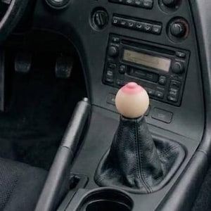 May include: The interior of a vehicle featuring a black dashboard, a steering wheel with a Toyota emblem, and a manual gear shifter topped with a spherical, skin-toned knob that has a small, raised, pinkish circular detail at the very top center.