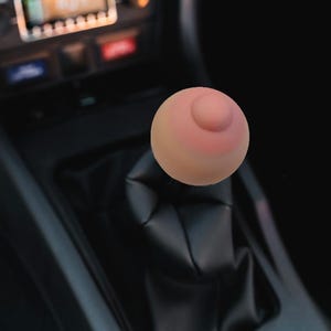 May include: A novelty shift knob shaped like a breast, featuring a pink nipple and areola. It's mounted on a black leather shift boot within a car's interior. The car's dashboard and radio are visible in the background.