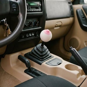 May include: A spherical gear shift knob featuring a pale white base and a prominent pink nipple-shaped protrusion at the top, mounted on a black rubber boot within the tan and black interior of a vehicle, positioned near the center console.