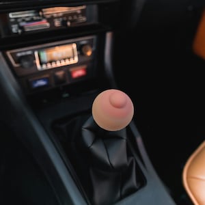 May include: A novelty shift knob shaped like a breast, featuring a pink nipple and areola. It's mounted on a black leather shift boot within a car's interior. The car's dashboard and radio are visible in the background.
