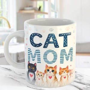 May include: White ceramic mug with the words "CAT MOM" in blue and white. The mug features illustrations of five cartoon cats with hearts and paw prints. The mug has a white handle and is on a white surface.