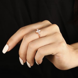 May include: Rose gold ring with a marquise-cut central stone and two smaller side stones. The band has a delicate, twisted design. The ring is shown on a hand with manicured nails against a black backdrop.