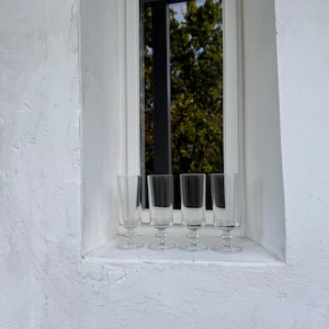 May include: Four clear, stemmed glasses sit on a white windowsill. The glasses have a slender, cylindrical bowl and a small, round base. The window frame is white, and the background shows green trees.