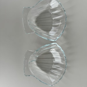 May include: Two clear glass seashell-shaped bowls. Each bowl has a scalloped edge and a handle on one side. The bowls are made of clear glass and have a ribbed interior design. The bowls are empty and sit on a white surface.