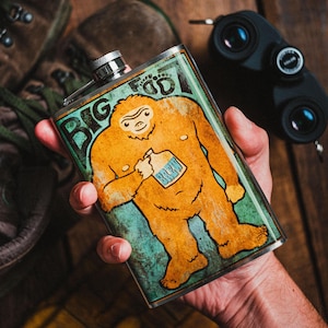 May include: Stainless steel flask with a Bigfoot graphic. The design features an orange Bigfoot holding a bottle labeled "Brew" against a teal and green background. The flask has a silver cap and is rectangular in shape.