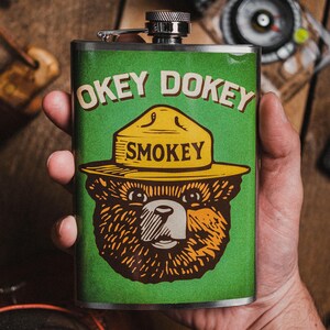 May include: A rectangular stainless steel flask with a green background featuring Smokey Bear wearing a yellow hat. The text "OKEY DOKEY SMOKEY" is printed on the front. The flask is held in a hand.