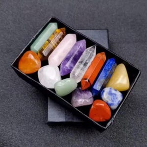 Peut inclure: Une collection de cristaux et de pierres précieuses colorées dans une boîte noire. Les cristaux varient en forme et en couleur, notamment orange, violet, transparent et bleu. La boîte est rectangulaire et posée sur une surface sombre.