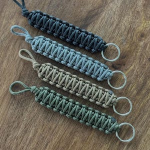 May include: Four woven paracord keychains in grey, blue, beige, and green. Each keychain has a loop at one end and a silver metal ring at the other. The keychains are displayed on a wooden surface, showcasing the craftsmanship.