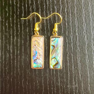 May include: A pair of gold-colored earrings with rectangular iridescent abalone shell inserts. The earrings have a gold-colored hook closure. The abalone shell displays a mix of blue, green, and gold colors. The earrings are displayed on a dark wooden surface.