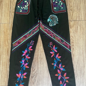 Op de afbeelding: Zwarte broek met een elastische tailleband met trekkoord, twee voorzakken met paarse en groene bloemenborduursels, een turquoise adelaarskop-afbeelding en levendige roze, blauwe en oranje bladpatronen geborduurd op de broekspijpen.