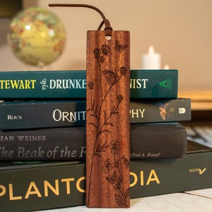 Op de afbeelding: Een bruine houten bladwijzer met een gegraveerd bloemen- en vlindermotief en een leren koord. De bladwijzer ligt op een stapel boeken. De boeken hebben titels als "The Drunkest", "Ornithology" en "Plantodia".