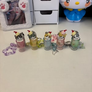 May include: Five miniature cat figurines in pastel-coloured cups. Each cat wears a small hat and sits on a colourful base. The figurines are linked by a beaded chain, with a star-shaped charm and a heart-shaped charm.