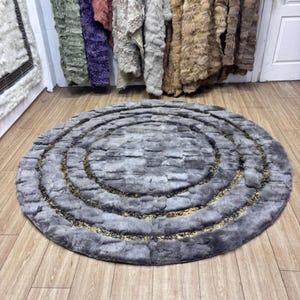 May include: A round, gray rug with concentric circles and gold accents. The rug is made of a soft, textured material and sits on a wooden floor. Various fur pelts hang in the background, suggesting a home decor theme.