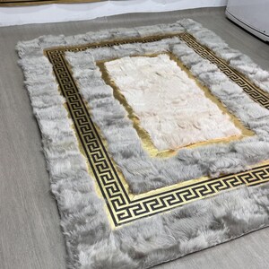May include: A rectangular area rug featuring a plush, light gray faux fur border and a central section of soft white faux fur. A gold-colored band with a black Greek key pattern separates the two fur textures, creating a distinct geometric frame.