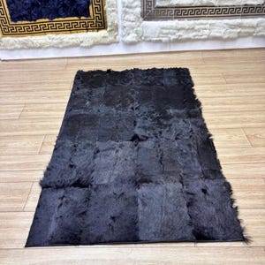 May include: A rectangular black fur rug, constructed from multiple patches. It is positioned on a light brown wooden floor. Other rugs with gold and white patterns are visible in the background.