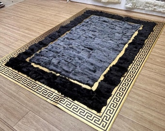 Puede incluir: Una alfombra rectangular con un borde negro y un diseño de llave griega dorada. El centro es de material gris texturizado, rodeado por un borde negro. La alfombra está sobre un suelo de madera.