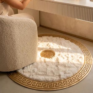 May include: A round white faux fur rug featuring a gold-colored Greek key border pattern and a matching circular gold-colored emblem in the center. The rug has a soft, textured pile and is positioned on a smooth floor surface.