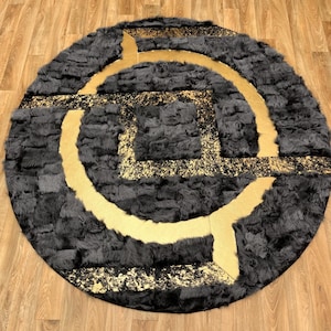 Puede incluir: Una alfombra circular con una textura de piel sintética en gris carbón profundo, contrastada por bandas geométricas lisas de color dorado y una sección cuadrada central con un acabado moteado en negro y oro de aspecto metálico.