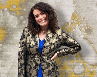May include: A woman wearing a black and gold floral patterned blazer with a blue dress underneath. She is standing in front of a textured wall with yellow accents.