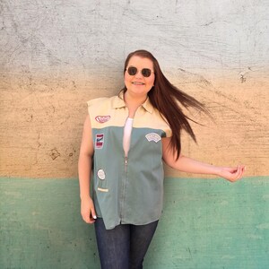 May include: A woman wearing a blue and yellow sleeveless jacket with patches and a zipper. She is wearing blue jeans and has brown hair. She is smiling and wearing sunglasses.