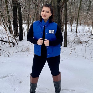 May include: A person wearing a blue quilted vest with a white patch, black long-sleeve shirt, black pants, and black and brown boots. The person is standing in a snowy forest.