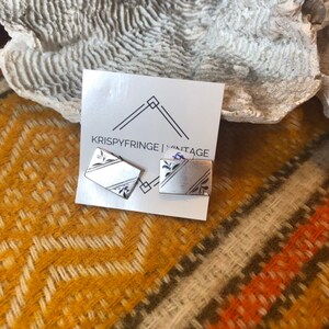 May include: A pair of silver cufflinks with a geometric design. The cufflinks are on a white card with the text "KRISPYFRINGE VINTAGE".