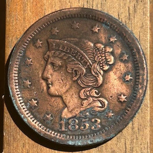 May include: A copper coin with a profile of a woman, the word "LIBERTY" above her head, and the date "1853" below. The coin has a detailed design with stars around the edge and a worn, aged appearance.