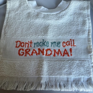May include: A white terry cloth bib featuring a central band of Aida fabric with the embroidered text "Don't make me call GRANDMA!" in orange and teal thread. The bib has a rounded neckline and a fringed bottom edge with a textured fabric finish.