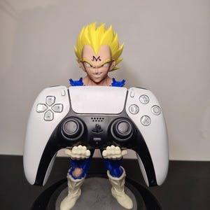 Peut inclure: Une figurine représentant un personnage aux cheveux en pointe avec un symbole M noir sur le front, vêtu d'une combinaison bleue et de bottes blanches, tenant une manette de jeu vidéo blanche et noire. Le personnage sourit largement.