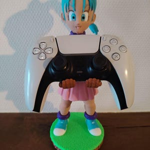 Op de afbeelding: Een beeldje van een personage met blauw haar en een roze jurk dat een wit en zwarte gamecontroller vasthoudt. Het beeldje staat op een groene basis met een bruine rand. De controller heeft knoppen en een touchpad.