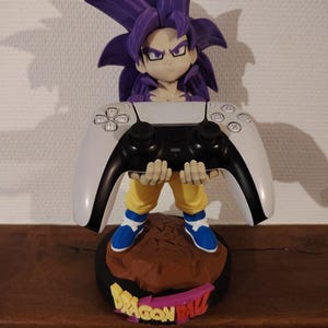 Peut inclure: Support de manette sur le thème de Dragon Ball Z. La figurine a des cheveux violets hérissés et un pantalon jaune, tenant une manette de jeu blanche et noire. La base est marron avec l'inscription "DRAGON BALL" en jaune et rose.
