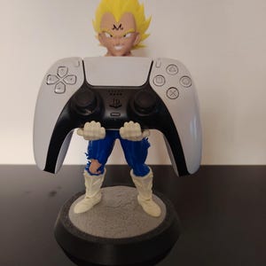 Puede incluir: Una figura de un personaje con cabello amarillo puntiagudo y una 'M' en la frente, sosteniendo un mando de PlayStation 5 blanco y negro. La figura está sobre una base circular gris y negra.