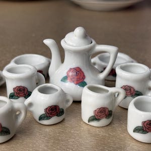 Peut inclure: Un service à thé miniature en porcelaine blanche comprenant une théière et plusieurs petites tasses et pichets. Chaque pièce est décorée d'une rose rouge et de feuilles vertes. L'ensemble est conçu pour une maison de poupée ou un présentoir décoratif.