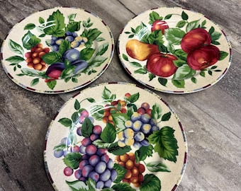 May include: Three decorative plates with fruit designs. Each plate features a different arrangement of grapes, apples, and plums, surrounded by green leaves. The plates have a cream-colored background with a red and white border.