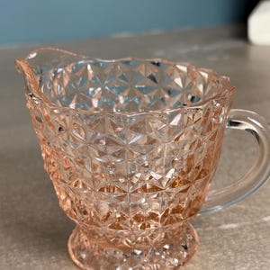 May include: A vintage pink glass creamer with a geometric pattern. The creamer has a handle and a spout for easy pouring. The glass is clear, allowing the contents to be visible. The creamer sits on a small, round base.