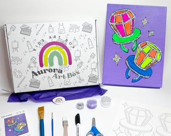 Könnte beinhalten: Eine Kinder-Kunstbox mit dem Text "Aurora Art Box" und einem Regenbogen-Logo. Die Box enthält Kunstzubehör, darunter Pinsel, Marker, Scheren und eine Leinwand mit Ring-Pop-Design. Ebenfalls enthalten sind Farbtöpfe und Wachsmalstifte.