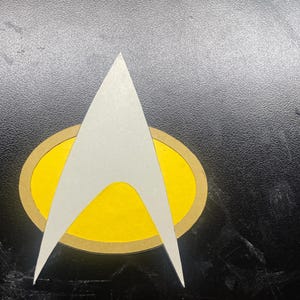 Peut inclure: Emblème Star Trek blanc et jaune, avec une forme de flèche stylisée sur un ovale jaune bordé de marron. L'emblème est posé sur un fond noir.