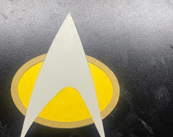 Puede incluir: Emblema de Star Trek blanco y amarillo, con una forma de flecha estilizada sobre un óvalo amarillo con borde marrón. El emblema está sobre un fondo negro.