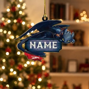 May include: A flat, hanging ornament featuring a dark blue dragon with large eyes and wings, positioned above a rectangular wooden-style sign with the word NAME printed in white capital letters, finished with a small red tail fin detail.