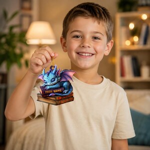May include: A flat, decorative ornament featuring a blue dragon with purple wings sleeping on a stack of two books. The top book has the text YOUR NAME printed in gold. The ornament includes a gold hanging cord for display purposes.