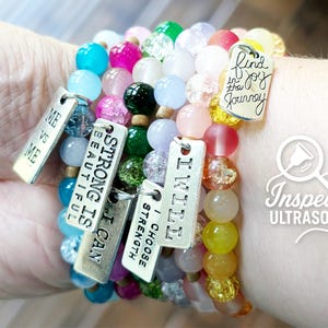 May include: A collection of colorful beaded bracelets with silver charms. The charms have inspirational phrases like "Strong is Beautiful" and "Find the Joy". The bracelets feature a variety of bead colors, including blue, pink, green, and yellow.