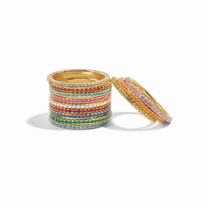 May include: A selection of gold-coloured rings, each featuring a band of colourful gemstones. The rings are stacked and arranged to display the variety of colours, including pink, blue, green, and yellow. The gemstones are small and evenly spaced.