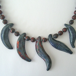 Peut inclure: Un collier artisanal composé de pendentifs incurvés bleu foncé avec des accents rouges, enfilés avec de petites perles marron et rouge foncé. Le collier a une esthétique tribale ou naturaliste.