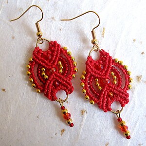 Puede incluir: Un par de pendientes de macramé rojos hechos a mano con detalles dorados. Los pendientes presentan intrincados patrones de nudos, pequeñas cuentas doradas y un grupo colgante de cuentas rojas y doradas. Los ganchos para las orejas son dorados.