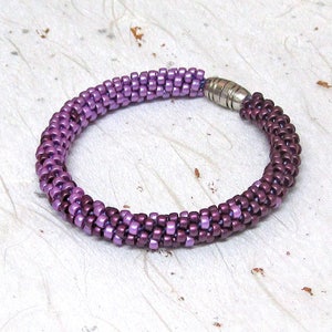 May include: A beaded bracelet in shades of purple, with a magnetic clasp. The bracelet is made of small, evenly spaced beads, creating a textured, cylindrical design. The color transitions from a lighter purple to a deeper shade.